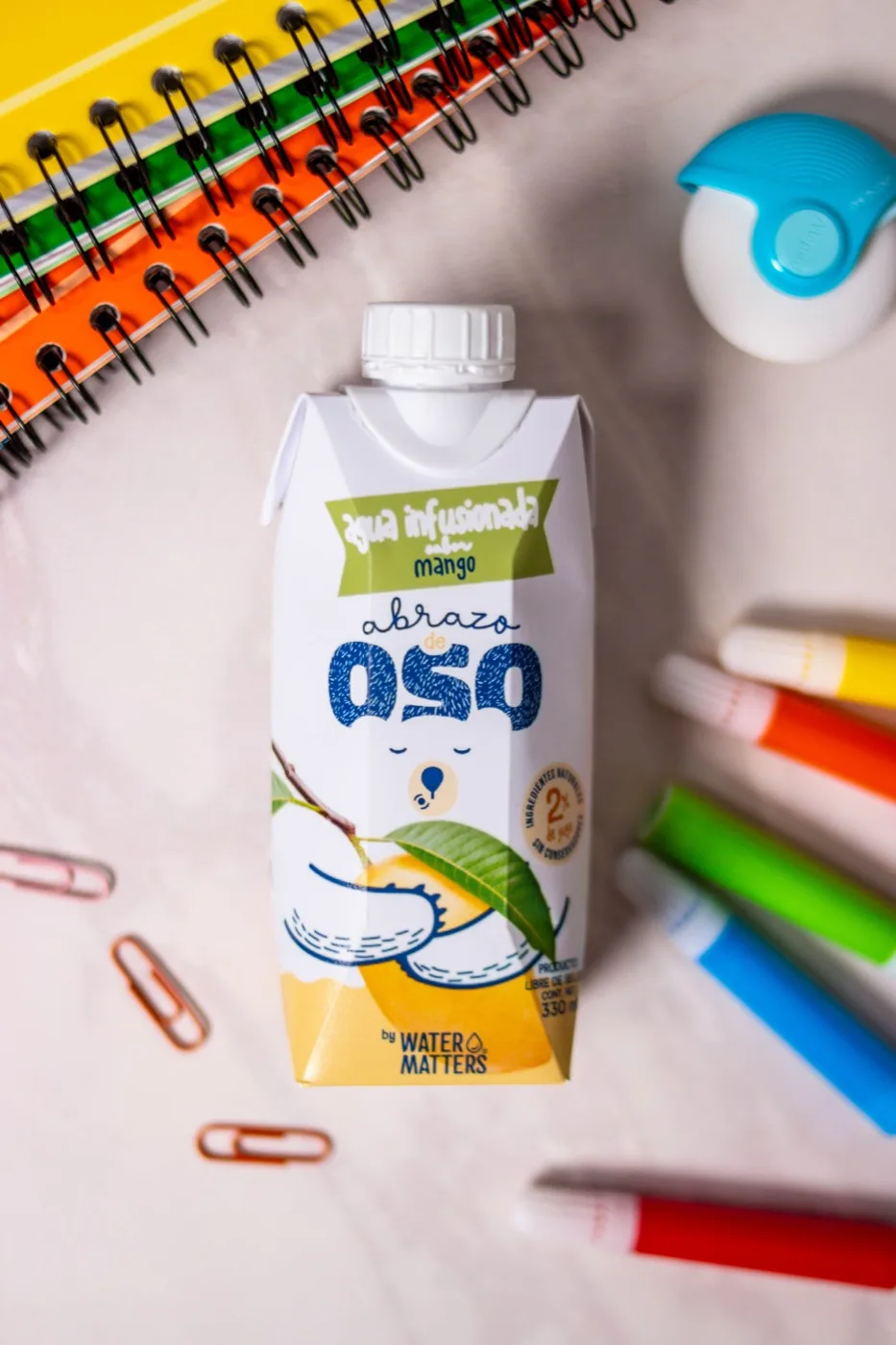 Envase de Abrazo de Oso sabor mango, con la firma «by Water Matters», entre útiles escolares