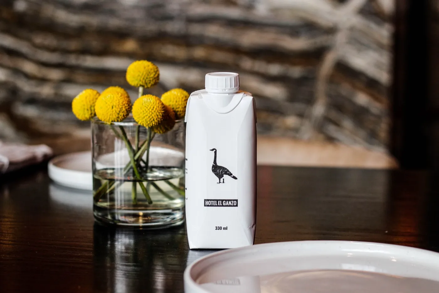 Un envase de Hotel El Ganzo junto a un vaso con flores amarillas, en la mesa del comedor