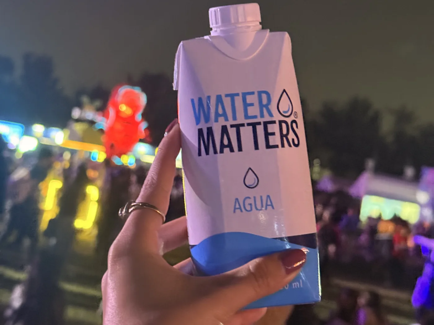 Un envase de Water Matters sostenido de noche en el Simifest, con el festival iluminado detrás